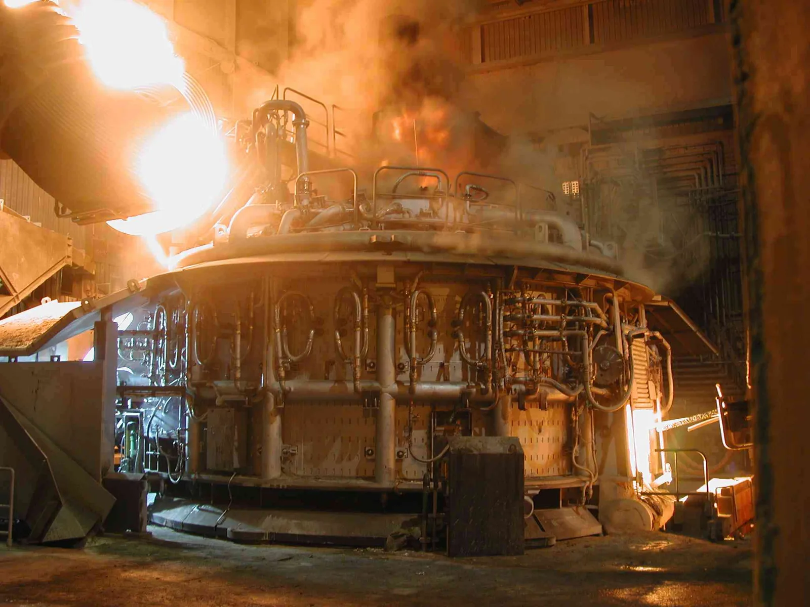 Electric arc furnace at a steelworks in operation, orange flames above the vessel and smoke in the hall