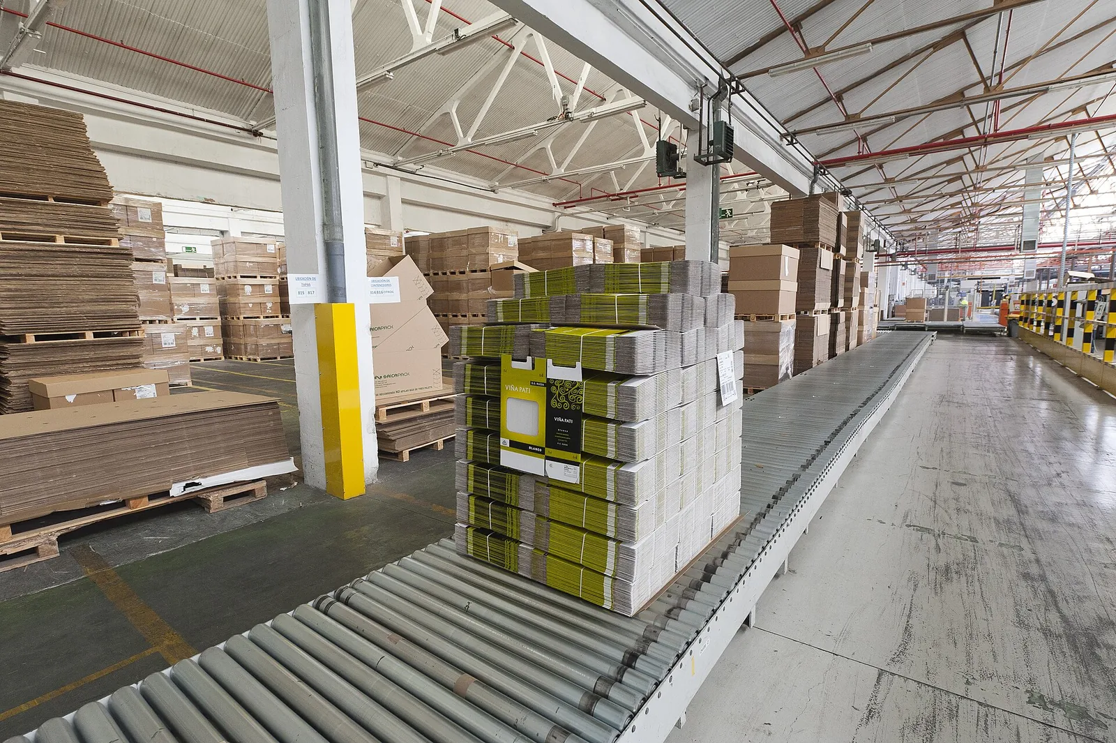 Pallet of folded, strapped corrugated cardboard boxes on a roller conveyor, in front of stacks of cardboard sheets in a factory hall