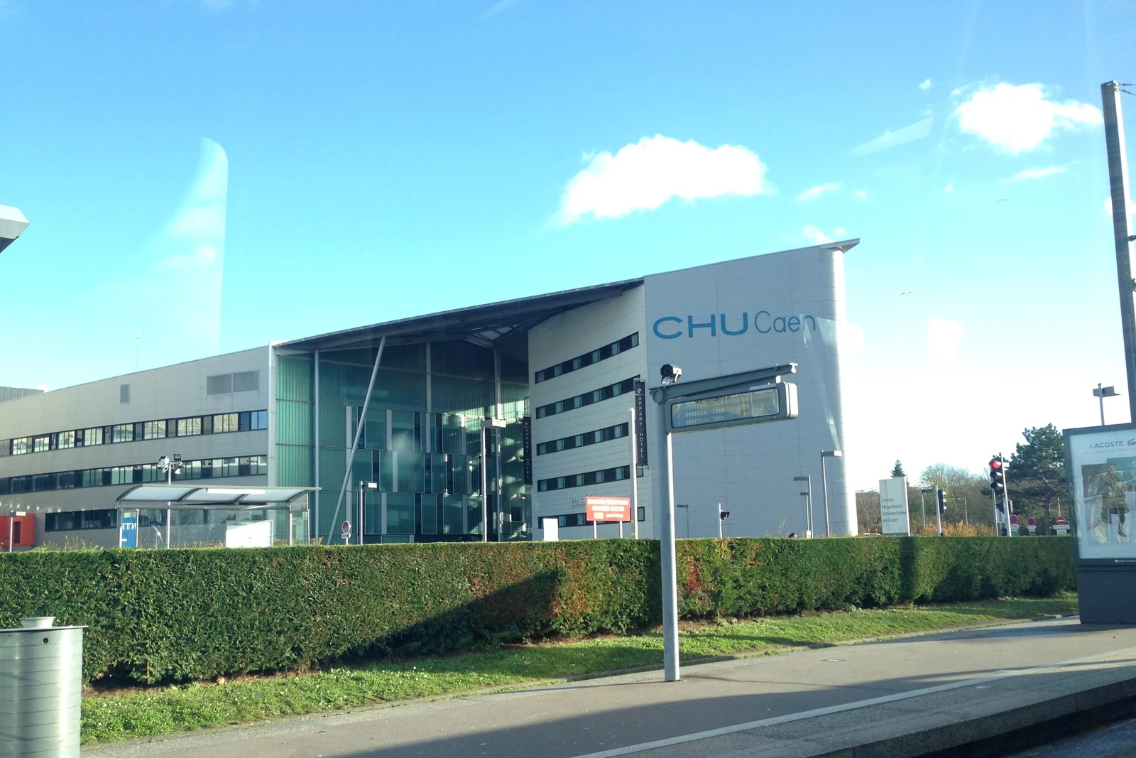 Glass façade of the Caen university hospital (CHU) under a blue sky, with the CHU Caen sign and a hedge in the foreground