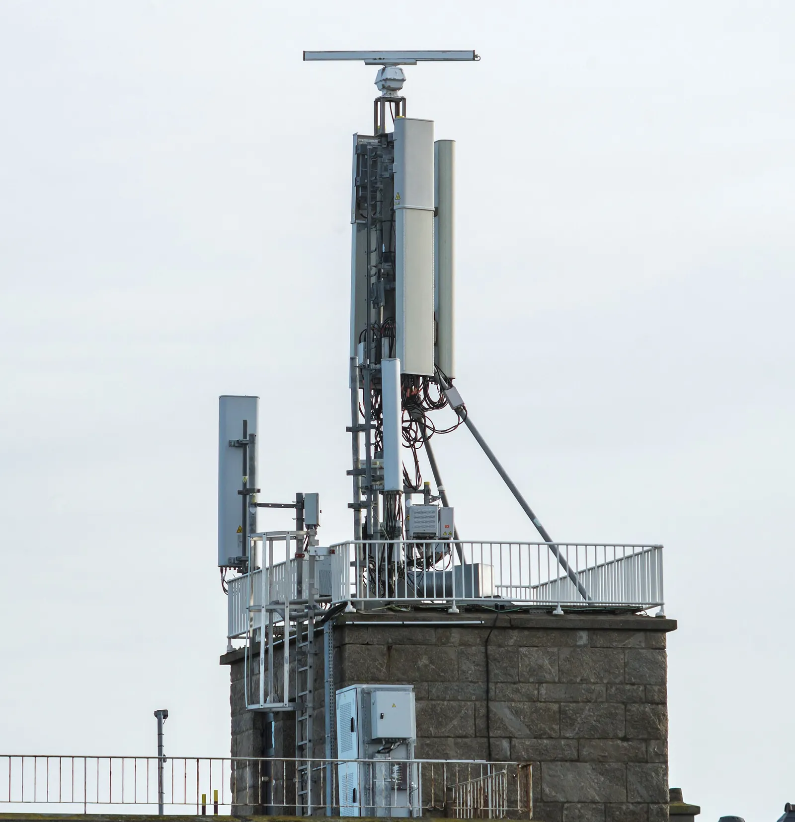 A mast of mobile phone antennas and its cables on top of a stone tower, behind a railing