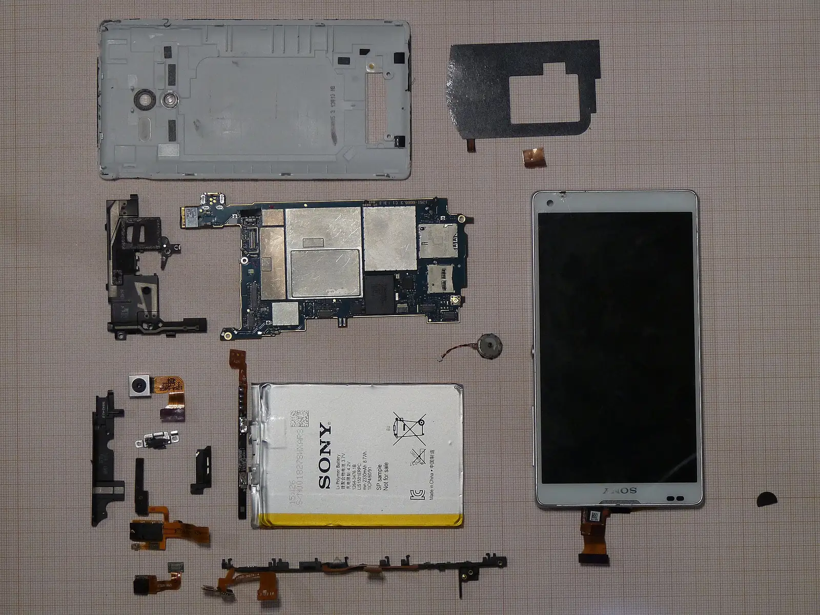 A disassembled smartphone, its parts laid flat on graph paper: casing, motherboard, battery, screen