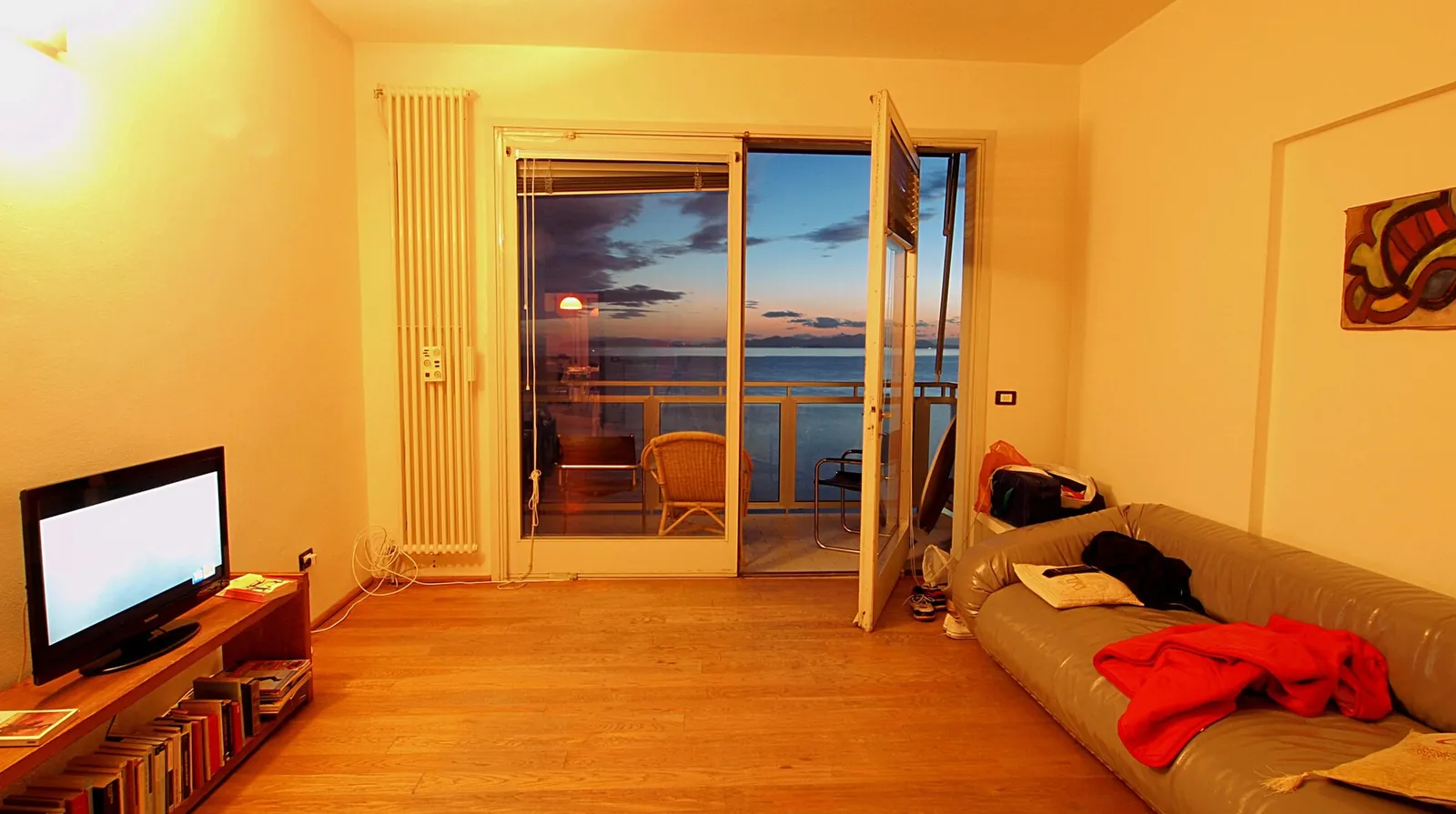 A living room at sunset, a television switched on on a low cabinet, a sofa and a French window open onto the sea